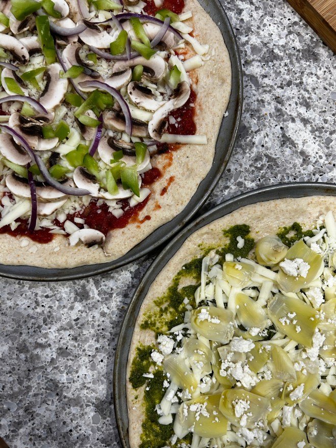 two pizzas on pans side by side at an skewed angle. top pizza had red sauce, mozzarella, green bell peppers and res onions. bottom pizza has pesto, artichoke, and crumbled feta